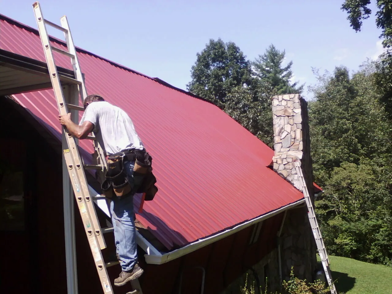 Roofer on a ladder beside a red metal roof and stone chimney — Soffit & Fascia Repair by Roofing Brothers in Denair